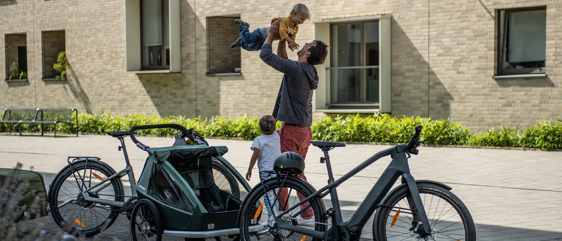 Two bikes in foreground, one with Croozer kid trailer and one with cargo trailer; father lifts son joyfully while another boy watches; house facade in background.